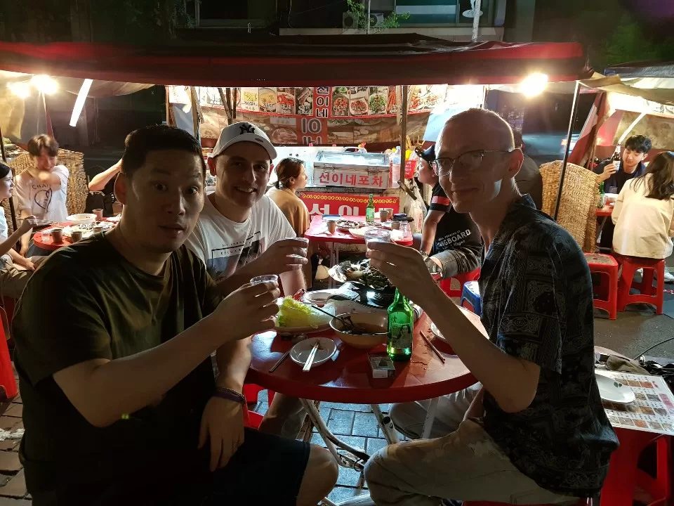 Trying street food in Seoul, cheering to the camera with soju glasses. 